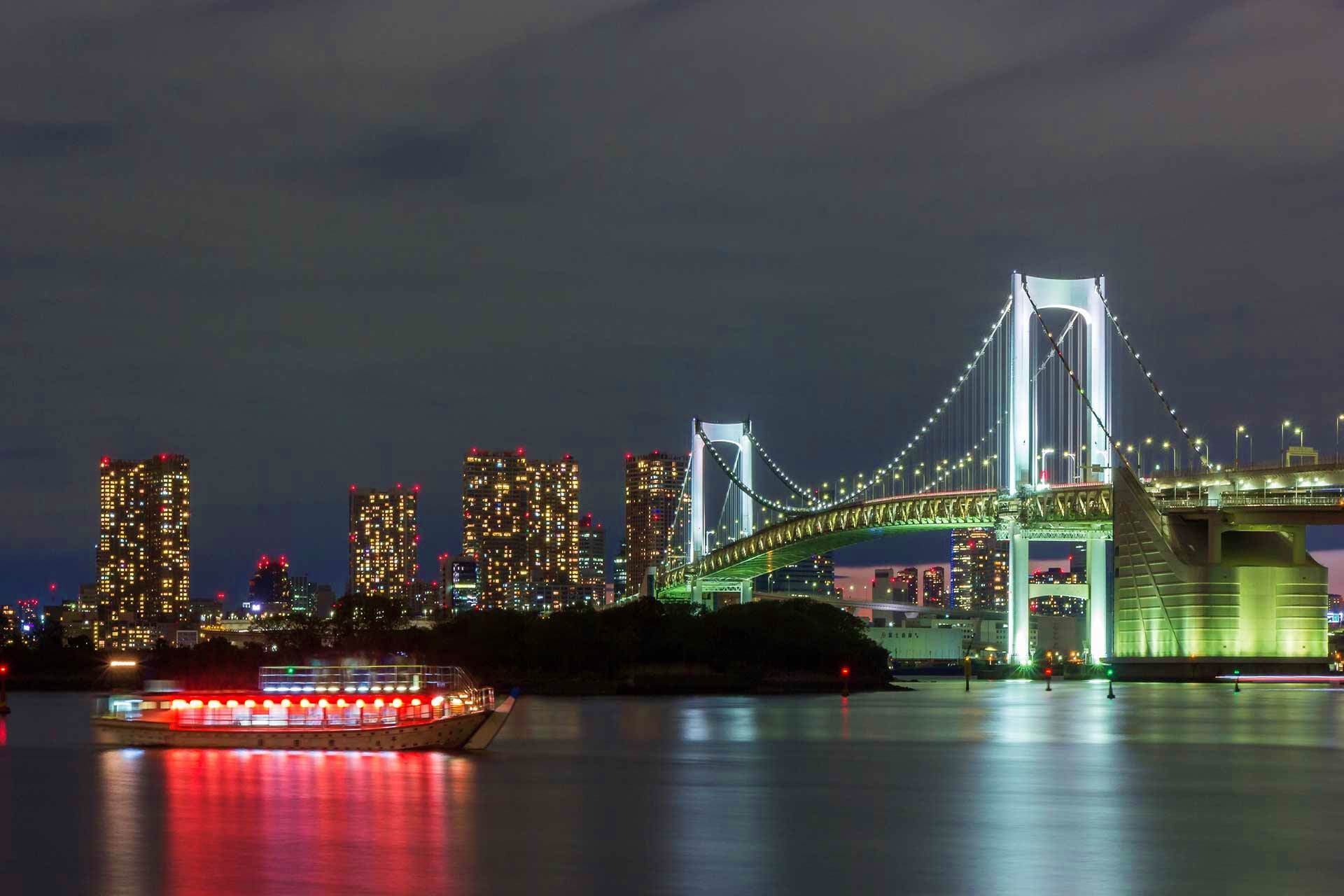Japan Visa Illuminated Rainbow Bridge spanning Tokyo Bay at night with lit passenger ferry boat and city skyline in background